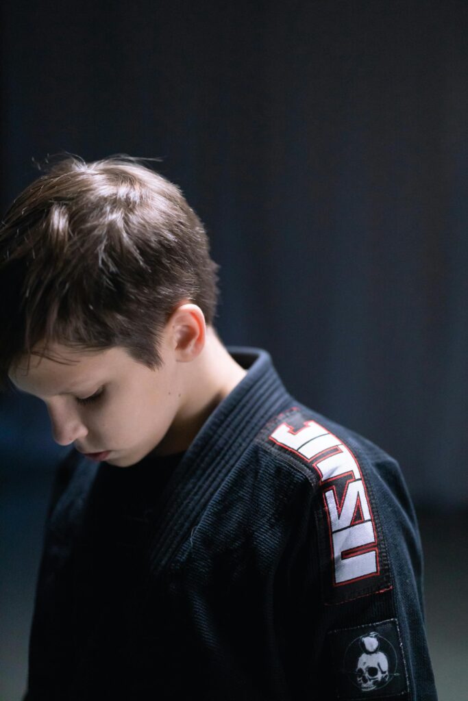 A young boy in a black jiu-jitsu gi looks down thoughtfully, captured indoors.